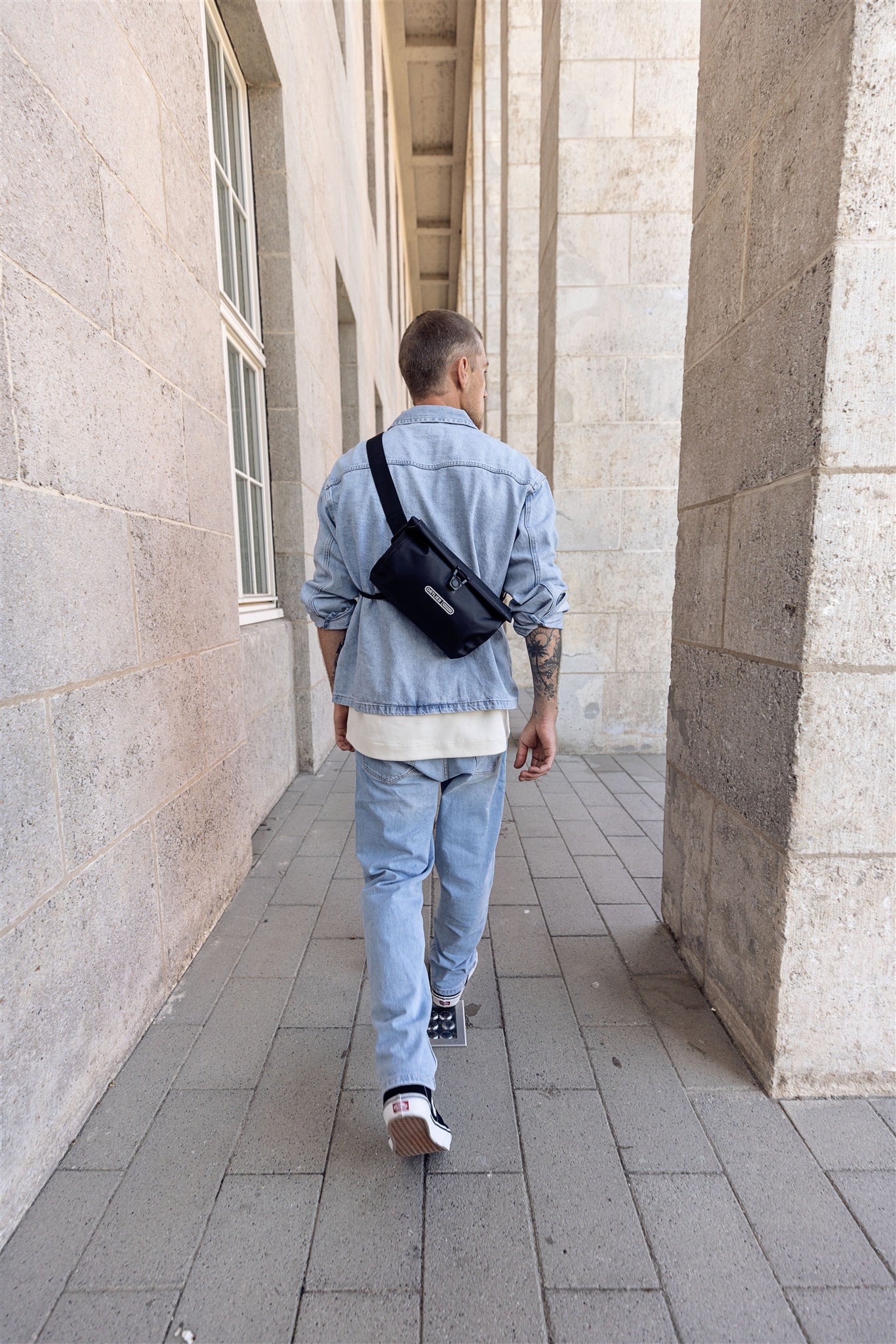 Achteraanzicht van man met zwarte ORTLIEB Velo-Sling Flex schoudertas met rolsluiting lopend door zuilengang.